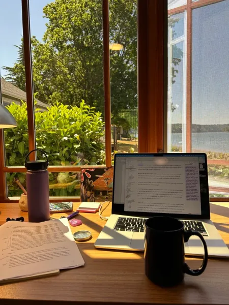 An open laptop computer on a desk next to a window along with note paper and pens, a coffee mug in the foreground, and a view of trees and water on a sunny day.